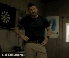 A Man In A Black Shirt And Khaki Pants Is Standing In Front Of A Dart Board In A Room . GIF
