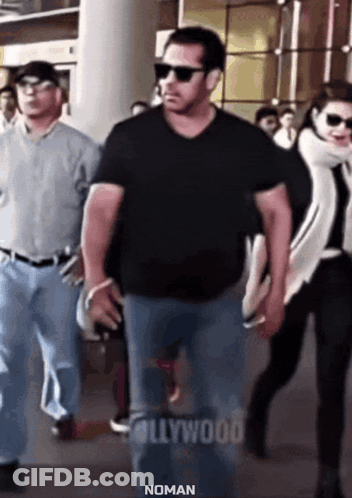 A Man In A Black Shirt And Sunglasses Is Walking With A Woman In A White Scarf Behind Him . GIF