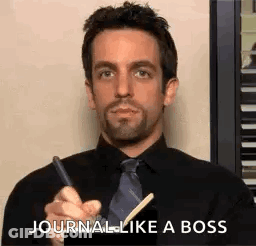 A Man In A Black Shirt And Tie Is Holding A Pen And Writing In A Notebook . GIF