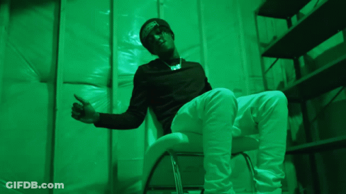 A Man In A Black Shirt And White Pants Is Sitting In A Green Room GIF