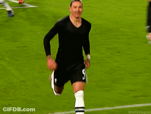 A Man In A Black Shirt And White Socks Stands On A Soccer Field Next To A Red Flag GIF