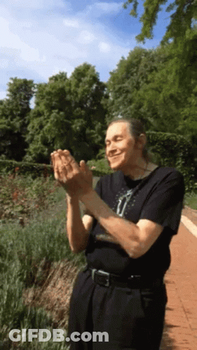 A Man In A Black Shirt Is Clapping His Hands In A Garden GIF