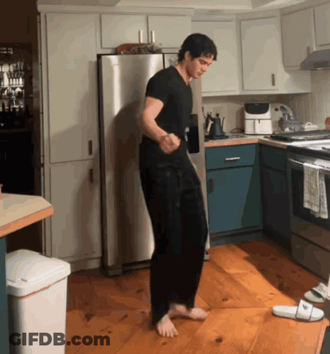 A Man In A Black Shirt Is Dancing In A Kitchen Next To A White Trash Can GIF