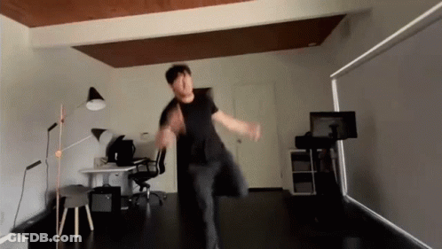 A Man In A Black Shirt Is Dancing In A Room With A Desk And Chair GIF