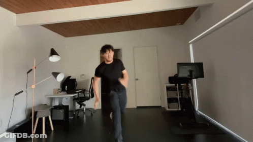 A Man In A Black Shirt Is Dancing In A Room With A Monitor On A Stand GIF
