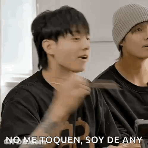 A Man In A Black Shirt Is Eating With Chopsticks While Another Man In A Beanie Looks On . GIF