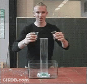 A Man In A Black Shirt Is Holding Two Shot Glasses In Front Of A Glass Container GIF