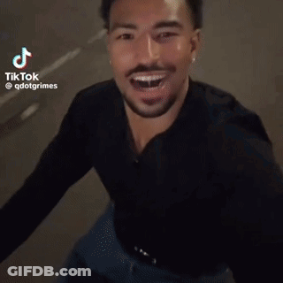 A Man In A Black Shirt Is Laughing And Smiling While Standing On A Street . GIF