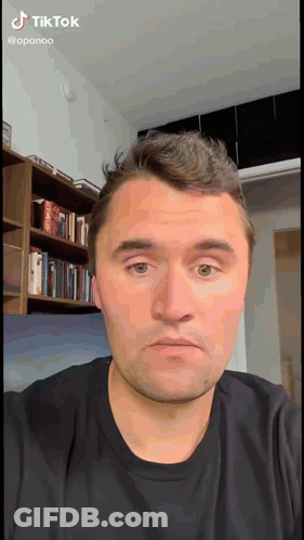 A Man In A Black Shirt Is Looking At The Camera With A Bookshelf In The Background GIF