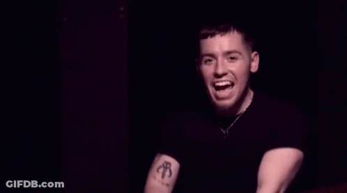 A Man In A Black Shirt Is Pointing At The Camera And Smiling In A Dark Room . GIF
