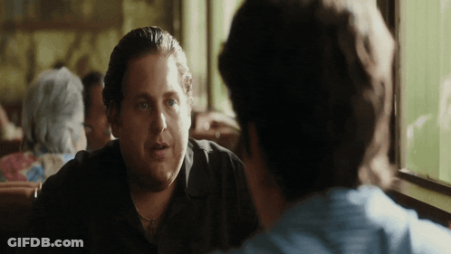 A Man In A Black Shirt Is Sitting At A Table Talking To Another Man GIF