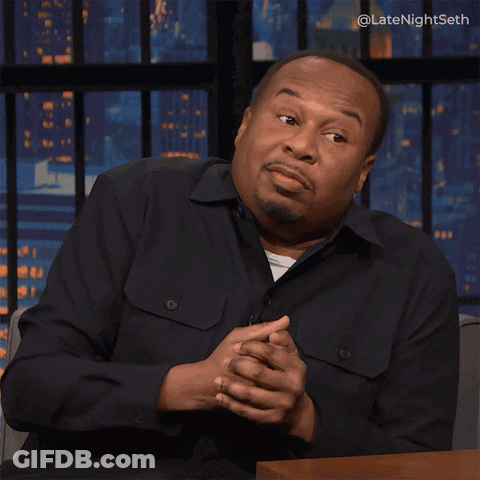 A Man In A Black Shirt Is Sitting At A Table With His Hands Outstretched In Front Of A Screen That Says Latenightseth GIF