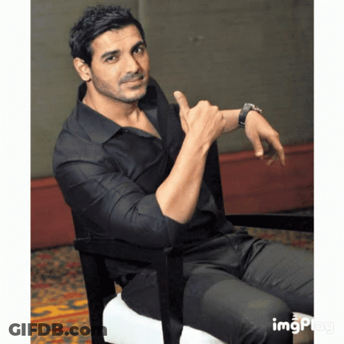 A Man In A Black Shirt Is Sitting In A Chair And Pointing At His Wrist GIF
