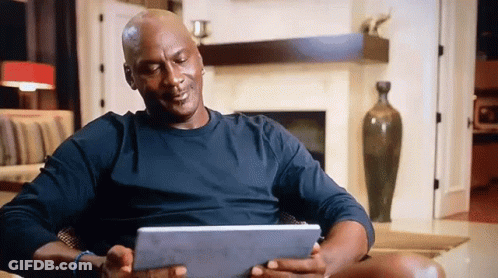 A Man In A Black Shirt Is Sitting In A Chair Holding A Tablet GIF