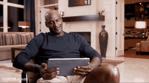 A Man In A Black Shirt Is Sitting In A Chair Using A Tablet GIF