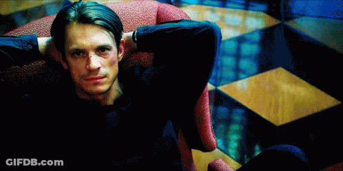 A Man In A Black Shirt Is Sitting In A Chair With His Hands Behind His Head GIF