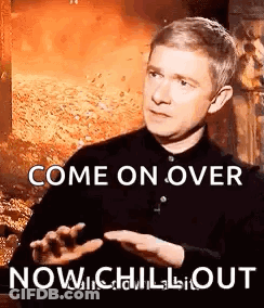A Man In A Black Shirt Is Sitting In Front Of A Fireplace And Talking To Someone While Holding His Hand Out . GIF