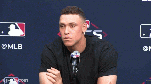 A Man In A Black Shirt Is Sitting In Front Of A Mlb Sign GIF