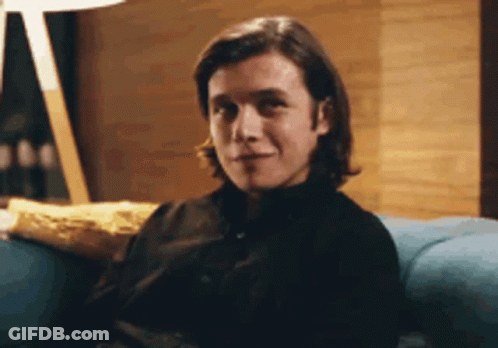 A Man In A Black Shirt Is Sitting On A Blue Couch Looking At The Camera . GIF