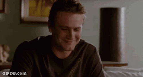 A Man In A Black Shirt Is Sitting On A Couch And Smiling . GIF