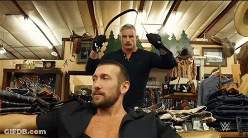 A Man In A Black Shirt Is Sitting On A Couch While Another Man Holds A Whip In A Store GIF