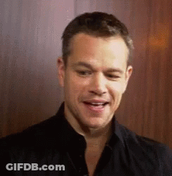 A Man In A Black Shirt Is Smiling And Looking At The Camera While Standing In Front Of A Wooden Wall . GIF