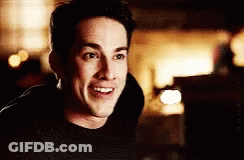 A Man In A Black Shirt Is Smiling In A Dark Room . GIF
