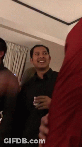 A Man In A Black Shirt Is Smiling In A Room With Other People GIF