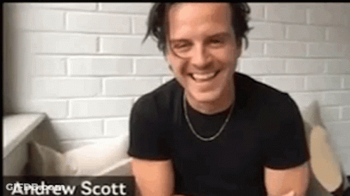 A Man In A Black Shirt Is Smiling In Front Of A Brick Wall . GIF