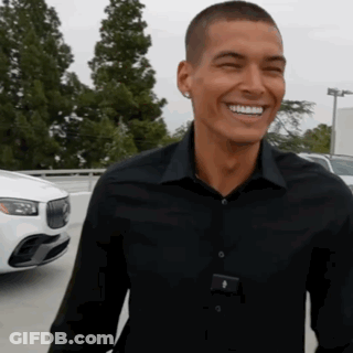 A Man In A Black Shirt Is Smiling In Front Of A White Car In A Parking Lot GIF