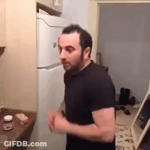 A Man In A Black Shirt Is Standing In A Kitchen Next To A Refrigerator . GIF