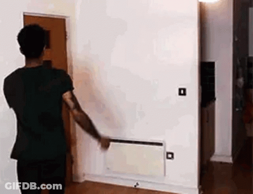 A Man In A Black Shirt Is Standing In A Living Room Looking At A Wall Heater . GIF