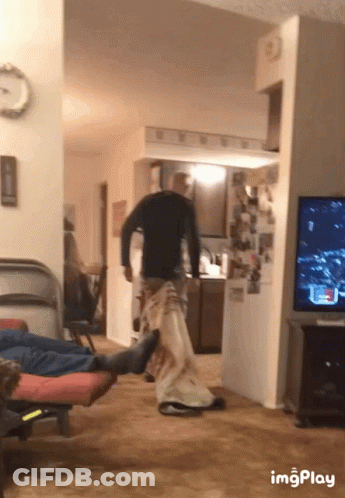 A Man In A Black Shirt Is Standing In A Living Room Next To A TV And A Clock GIF