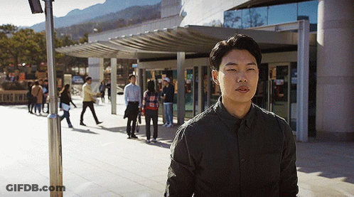 A Man In A Black Shirt Is Standing In Front Of A Building With People Walking In The Background GIF