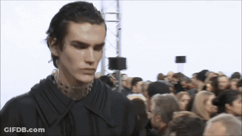 A Man In A Black Shirt Is Standing In Front Of A Crowd Of People GIF