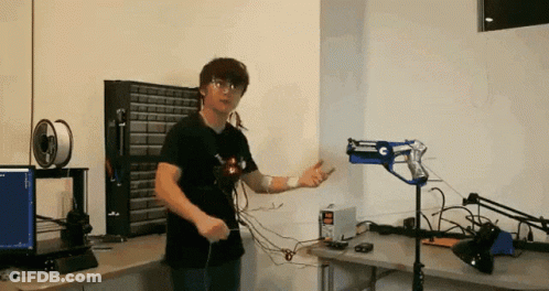 A Man In A Black Shirt Is Standing In Front Of A Desk With A Computer And A Gun On It GIF