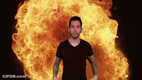 A Man In A Black Shirt Is Standing In Front Of A Large Explosion Of Flames GIF