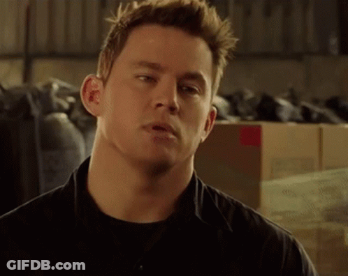 A Man In A Black Shirt Is Standing In Front Of A Pile Of Boxes And Looking At The Camera . GIF