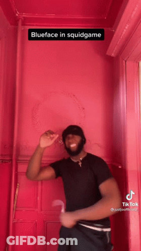 A Man In A Black Shirt Is Standing In Front Of A Pink Wall With The Caption Blueface In Squidgame . GIF