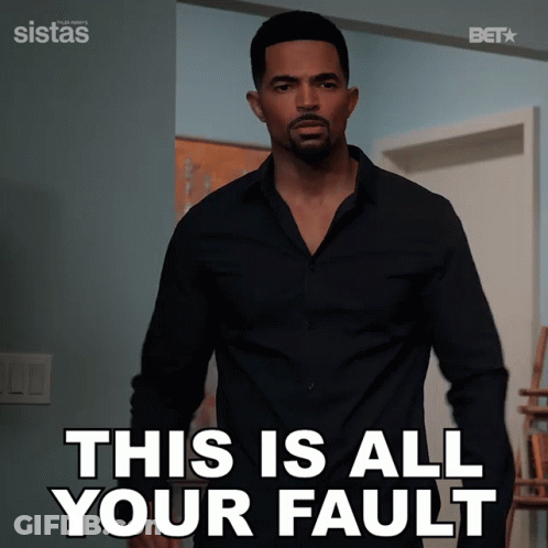 A Man In A Black Shirt Is Standing In Front Of A Sign That Says This Is All Your Fault GIF
