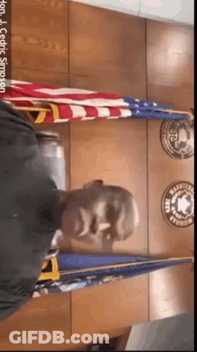 A Man In A Black Shirt Is Standing In Front Of A Wall With Flags And Badges On It .. GIF