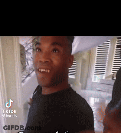 A Man In A Black Shirt Is Standing In Front Of A Window With Blinds . GIF