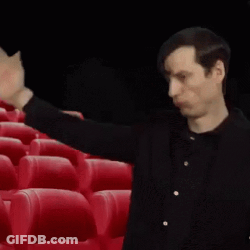 A Man In A Black Shirt Is Standing In Front Of Red Seats In A Theater . GIF