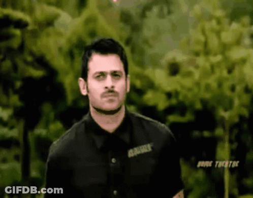 A Man In A Black Shirt Is Standing In Front Of Some Trees . GIF