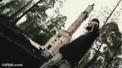 A Man In A Black Shirt Is Standing In The Woods In Front Of A Statue . GIF