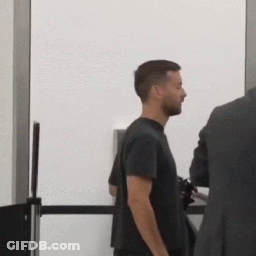 A Man In A Black Shirt Is Standing Next To A Man In A Suit At A Security Checkpoint . GIF
