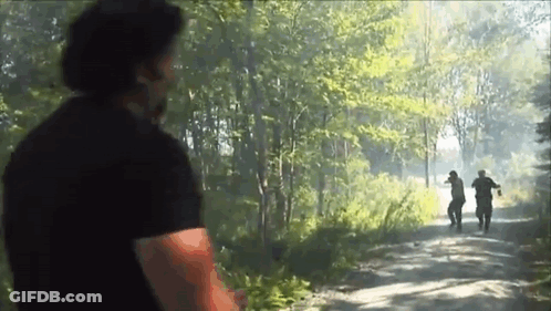 A Man In A Black Shirt Is Standing On A Dirt Road In The Woods Looking At Two Men Walking Down The Road . GIF
