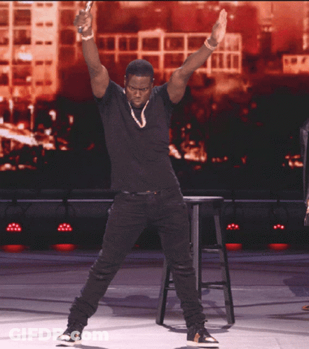 A Man In A Black Shirt Is Standing On A Stage With His Hands In The Air GIF