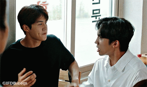 A Man In A Black Shirt Is Talking To Another Man In A White Shirt While Sitting In Front Of A Window . GIF