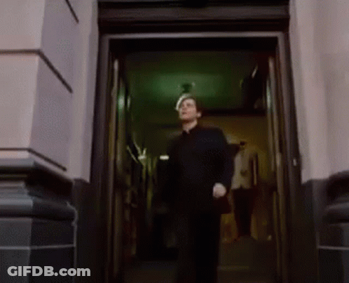 A Man In A Black Shirt Is Walking Out Of A Doorway . GIF